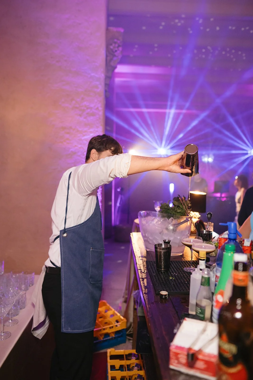 Bartender pouring a drink at a bar with colorful stage lights and DJ in the background, with various bottles and glasses on the counter.