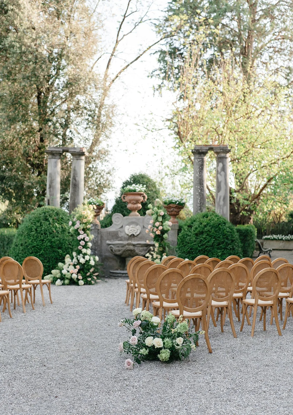 Outdoor wedding ceremony setup with wooden chairs arranged in rows, floral decorations, and a stone fountain with potted plants and trees in the background.