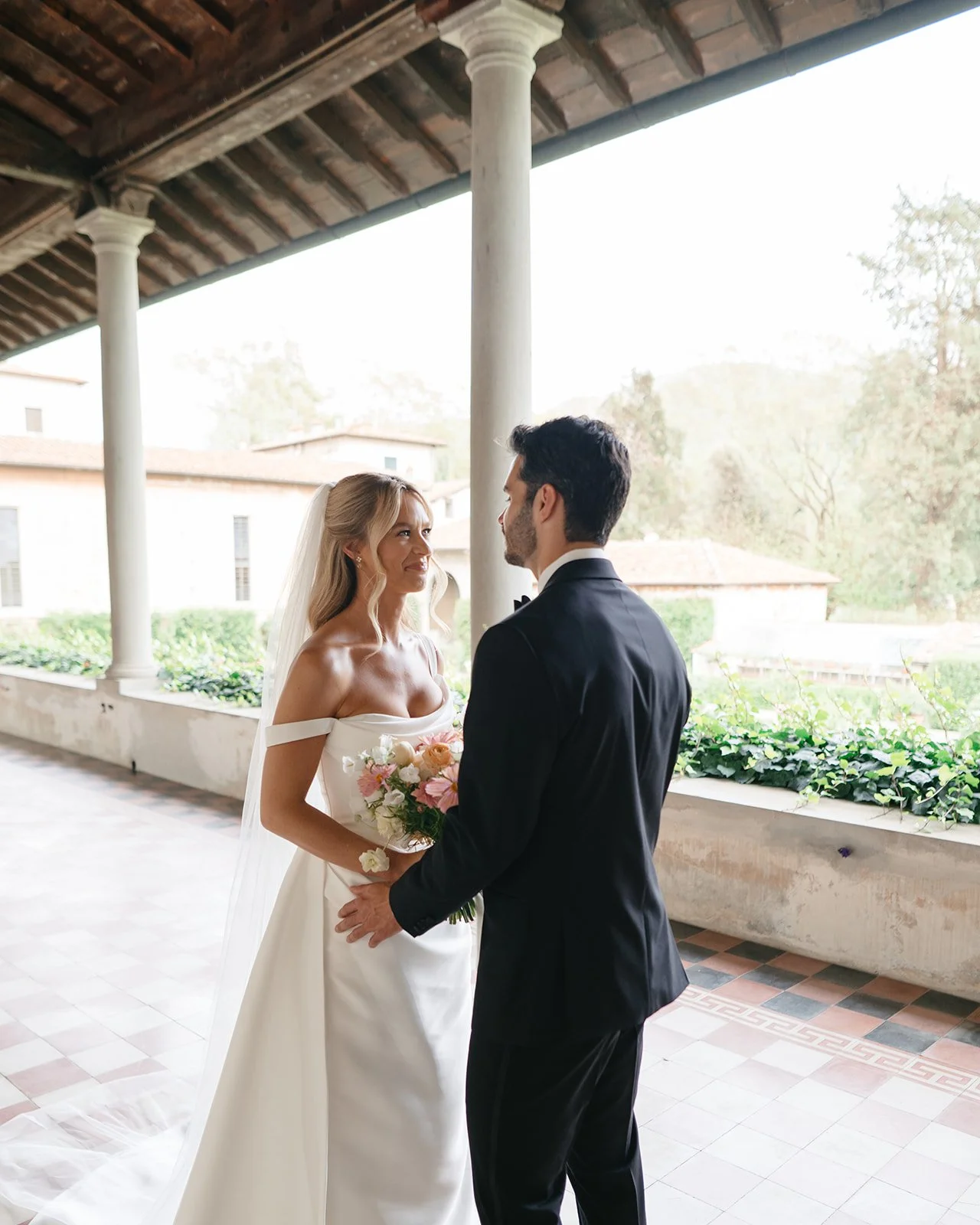 Bride and groom standing face-to-face on a covered outdoor walkway, with the bride holding a bouquet of flowers, during their wedding ceremony.