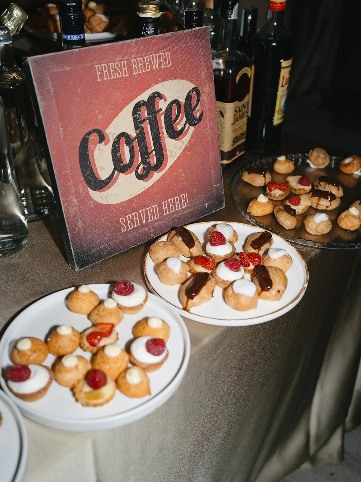 Assorted mini desserts on white plates, topped with strawberries, raspberries, and whipped cream, next to a vintage-style sign that says "Fresh Brewed Coffee Served Here," with bottles of alcohol and a plate of pastry in the background.