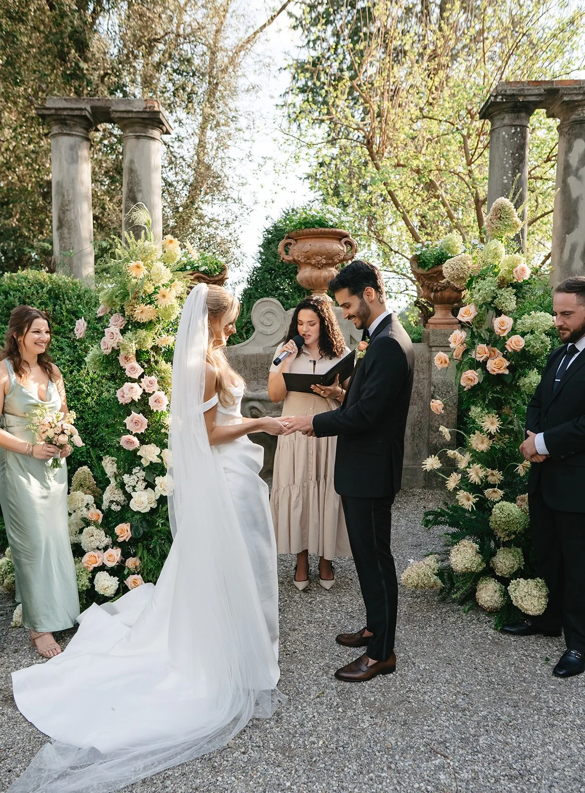 A wedding ceremony outdoors with a bride and groom exchanging vows, surrounded by bridesmaids, groomsmen, and floral decorations.