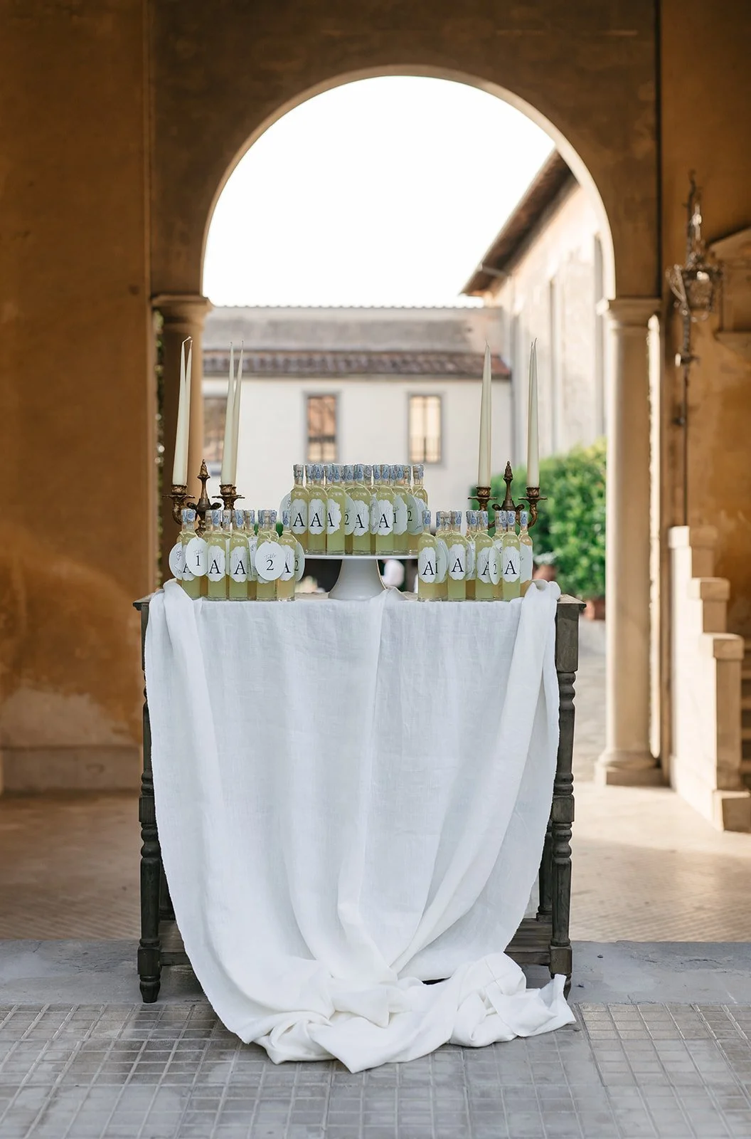 A table with white drapery features bottles with labels marked 'A1' and 'A2', topped with tall white candles, set in an elegant outdoor setting under an archway.