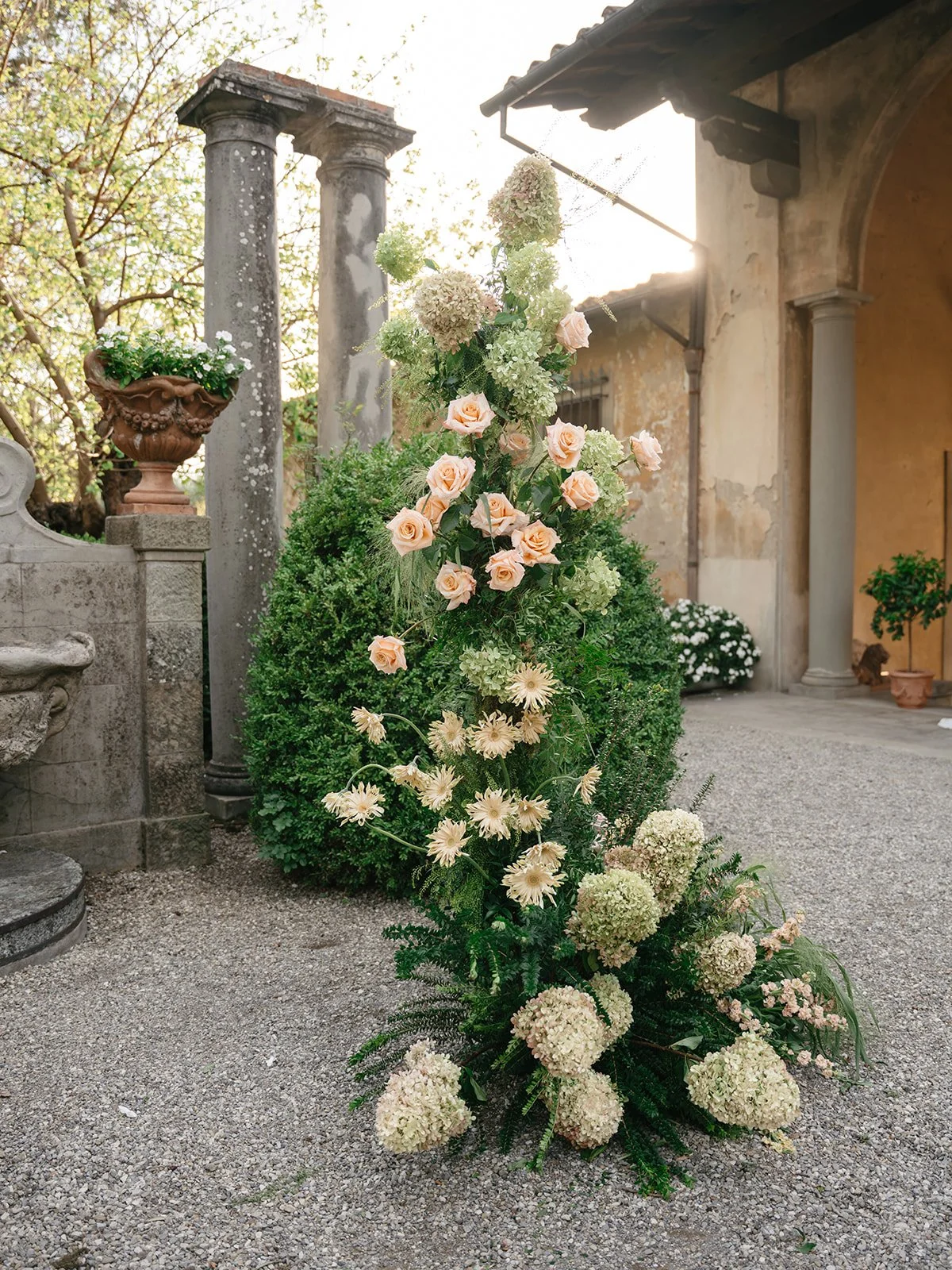 A floral arrangement with pink roses, white and green hydrangeas, and white gerbera daisies, set against an outdoor backdrop of stone columns, greenery, and an old building.
