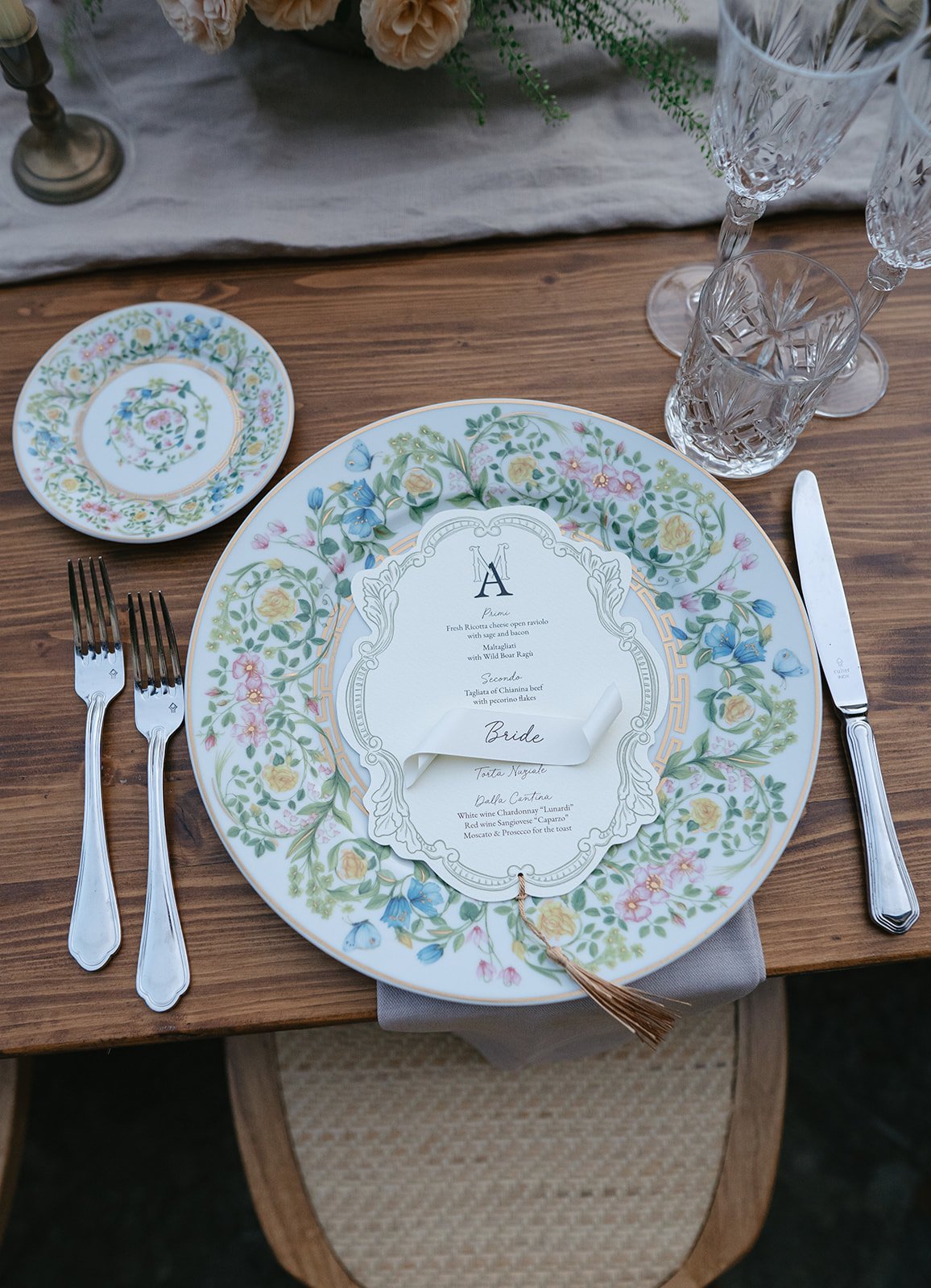 Elegant dinner table setting with floral patterned plates, a printed menu card, silverware, and crystal glassware, on a wooden table with a cloth napkin.