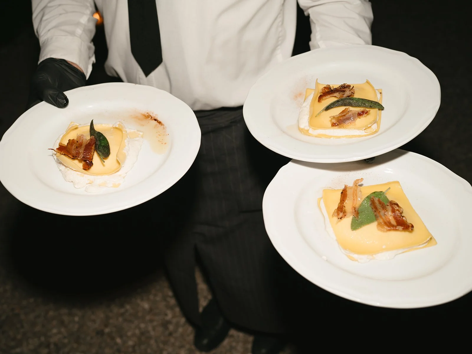 Waiter in white shirt and black gloves holding three white plates with slices of cheese, bacon, and sage leaves.