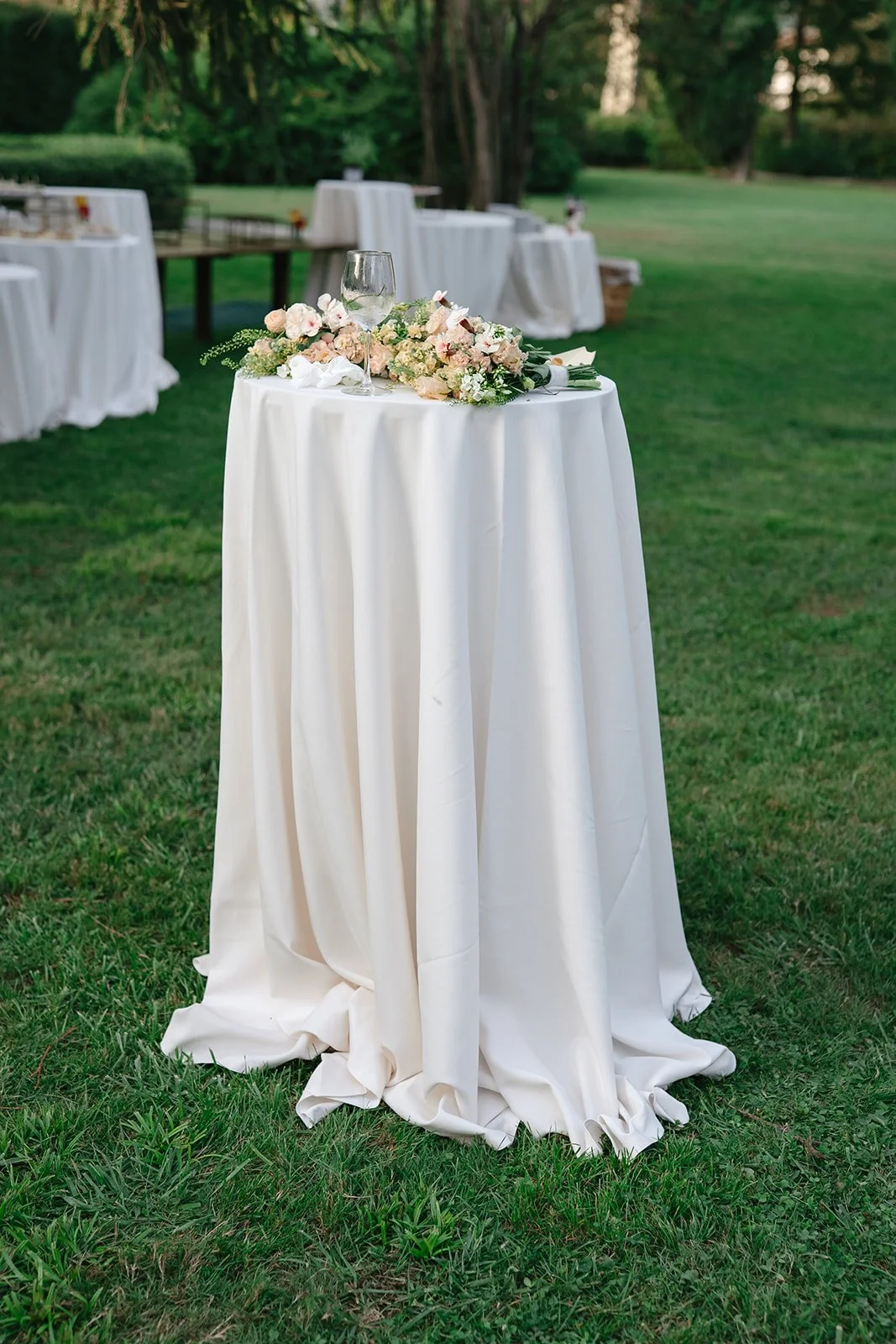 Round table with white tablecloth, decorated with a floral arrangement and a wine glass, set outdoors on grass with other tables in the background.