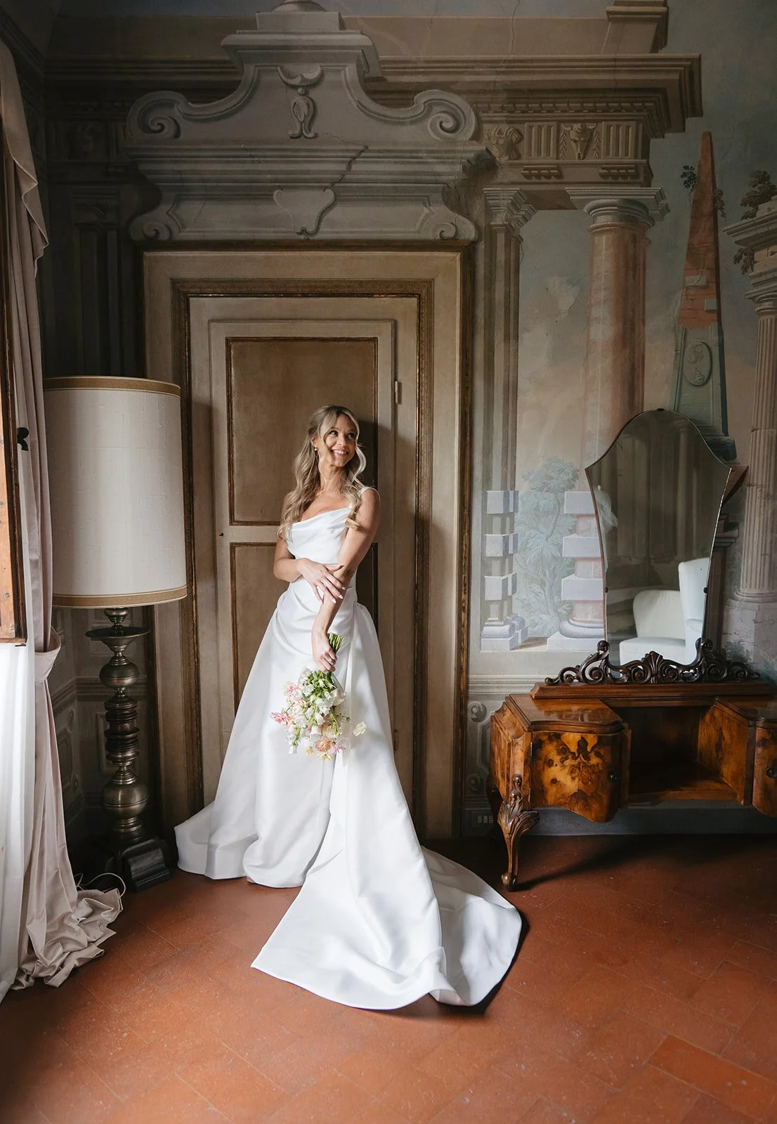 A woman in a white wedding dress holding a bouquet of flowers, standing in a room with painted walls and antique furniture, smiling and looking to the side.