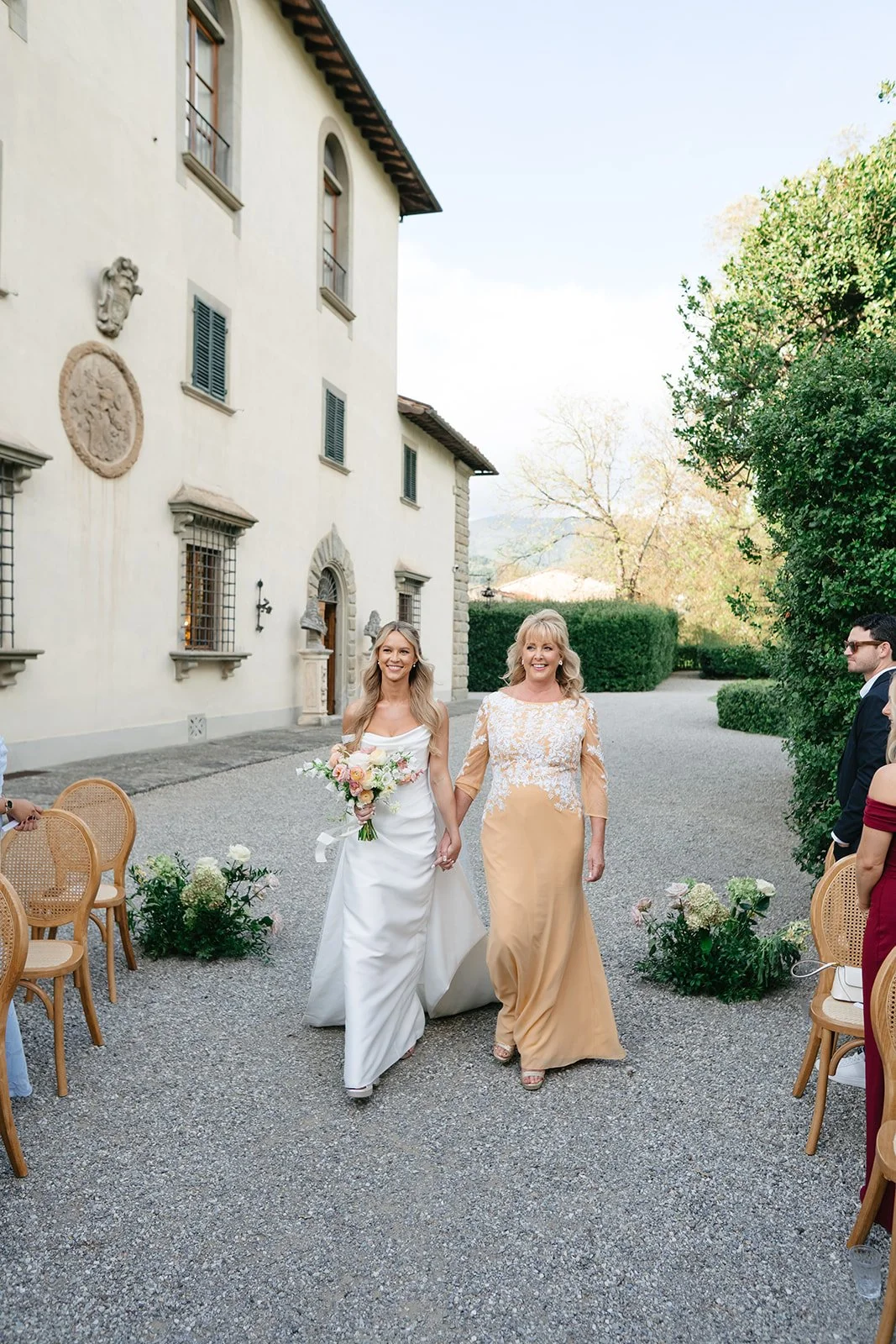 A bride and a woman walking hand in hand at an outdoor wedding ceremony, with guests on sides, in front of a large white building with historical architectural details, trees, and a gravel pathway.