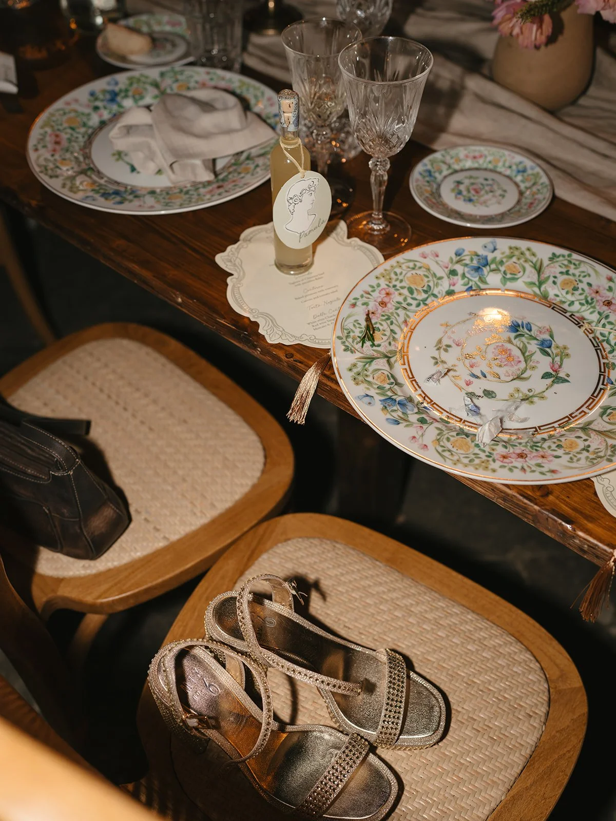 An elegantly set dining table with floral-patterned plates, glasses, a small bottle of drink, and a menu card. Two wooden chairs with woven seats are positioned in front of the table, one of which has a pair of gold glittering high-heeled sandals on 