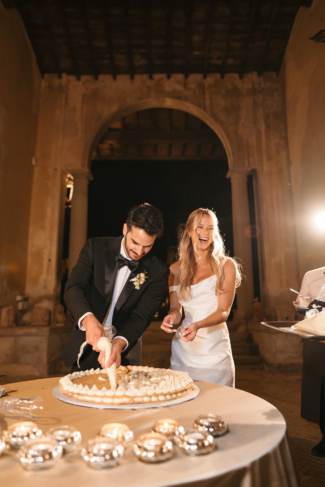 A newlywed couple cutting a wedding cake together at their reception, both smiling and laughing.