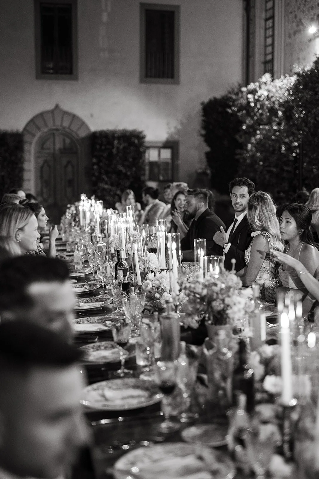 Guests seated at a long outdoor dinner table decorated with flowers and candles during evening.