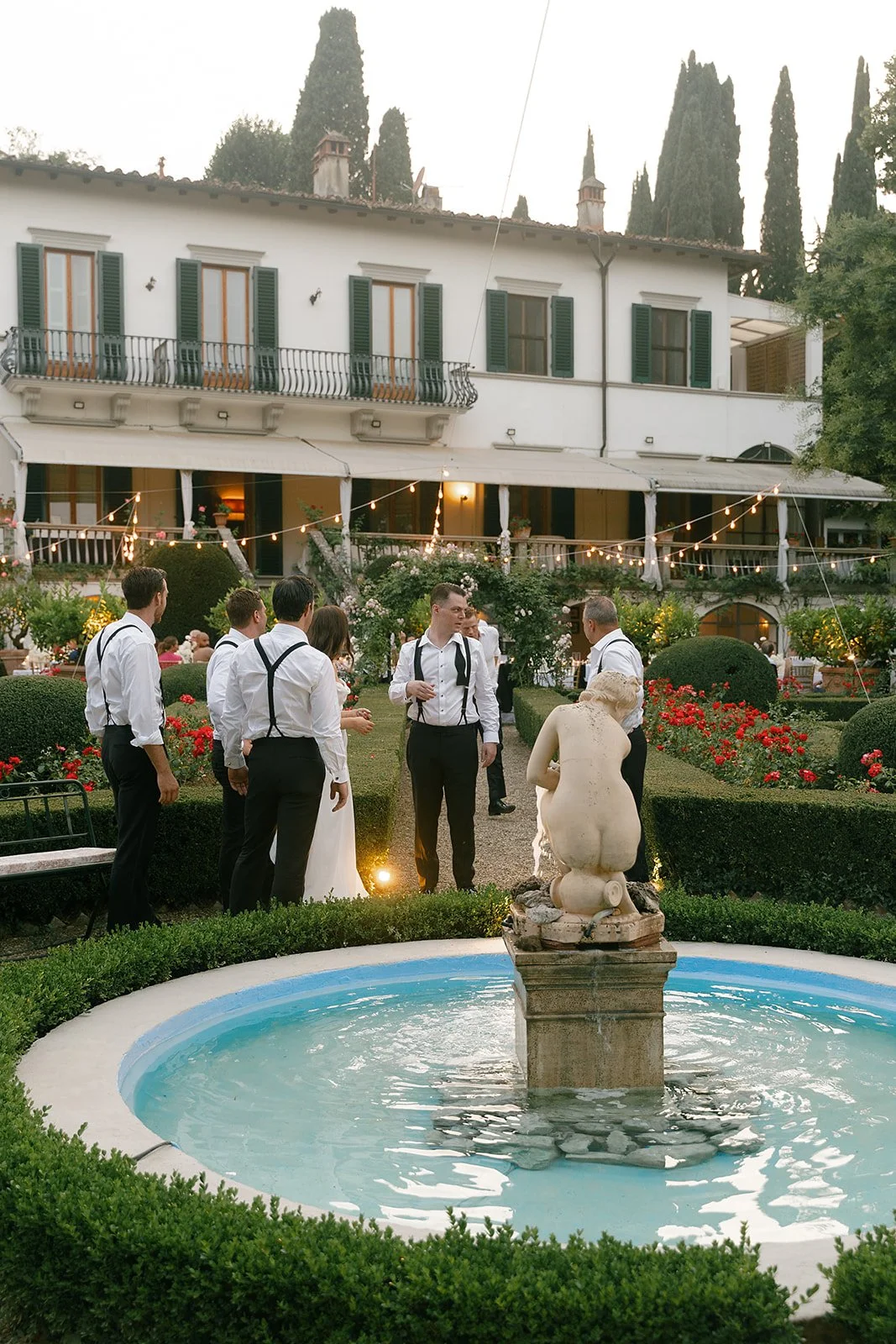 Group of people dressed in formal attire gathered in a garden with a fountain, in front of a white building with a terrace decorated with string lights.