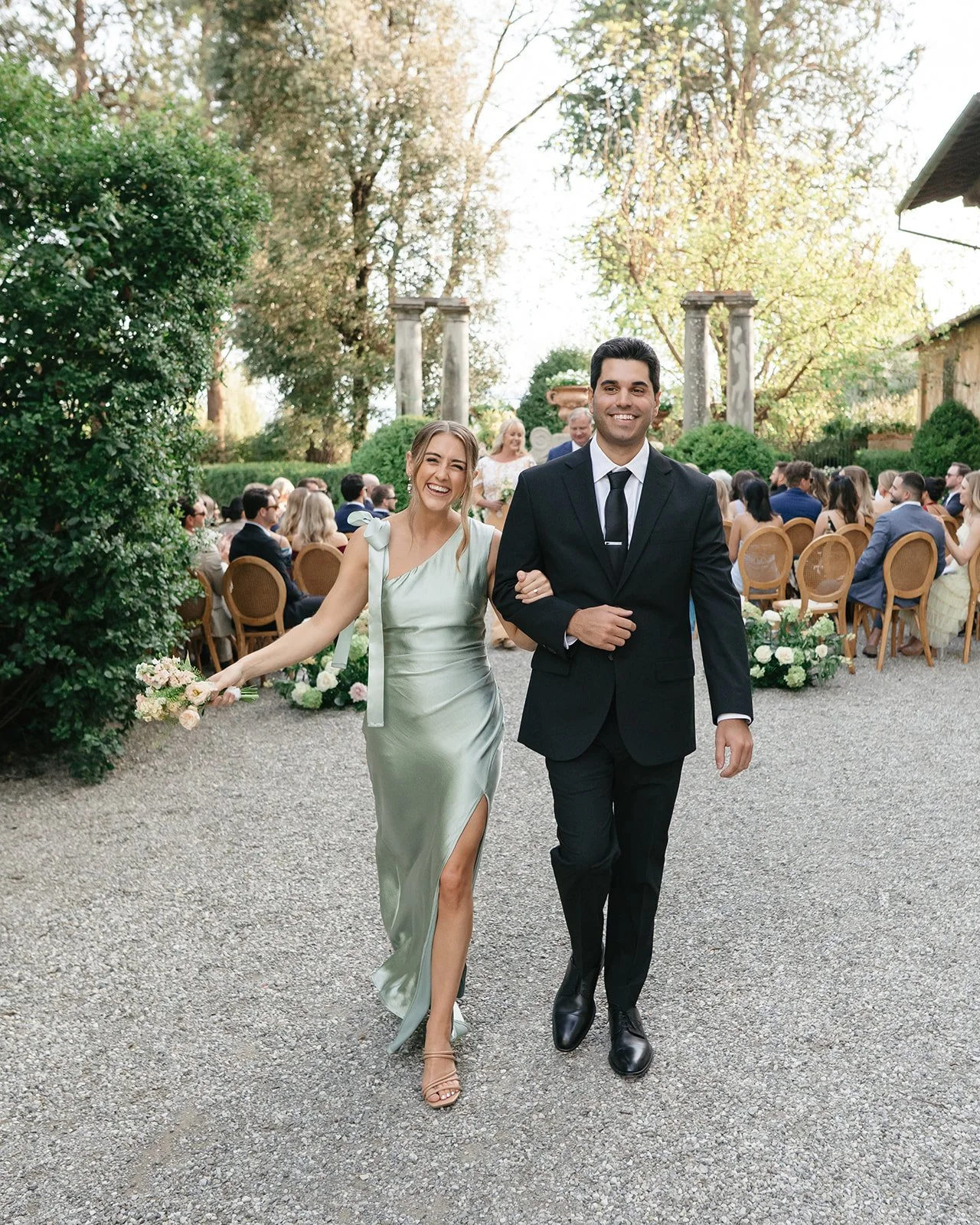 A smiling woman in a silver dress walking arm-in-arm with a smiling man in a black suit at an outdoor wedding ceremony with seated guests and decorative greenery in the background.