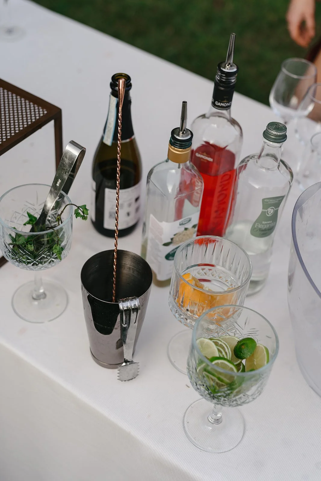 A table with various bottles of alcohol, cocktail glasses, and garnishes including lime and mint leaves at an outdoor setting.