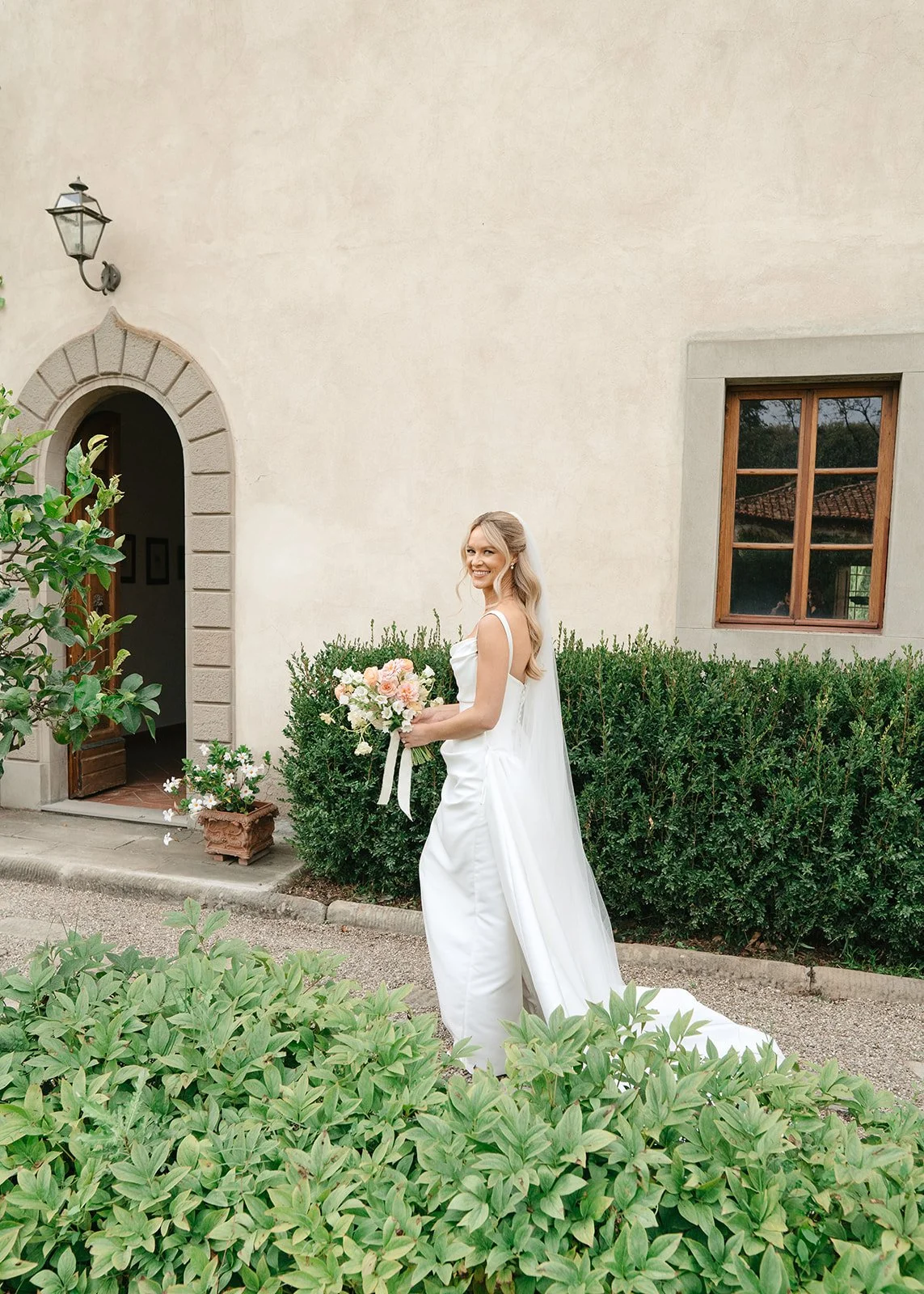 A bride in a white wedding gown holding a bouquet of pink and white flowers, standing outdoors in front of a beige building with a window and bushes.