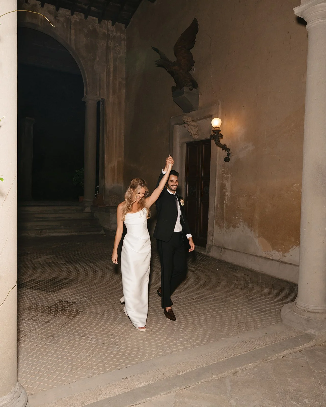 A bride and groom walking hand in hand outdoors at night, smiling and celebrating, with the groom raising their joined hands in victory.