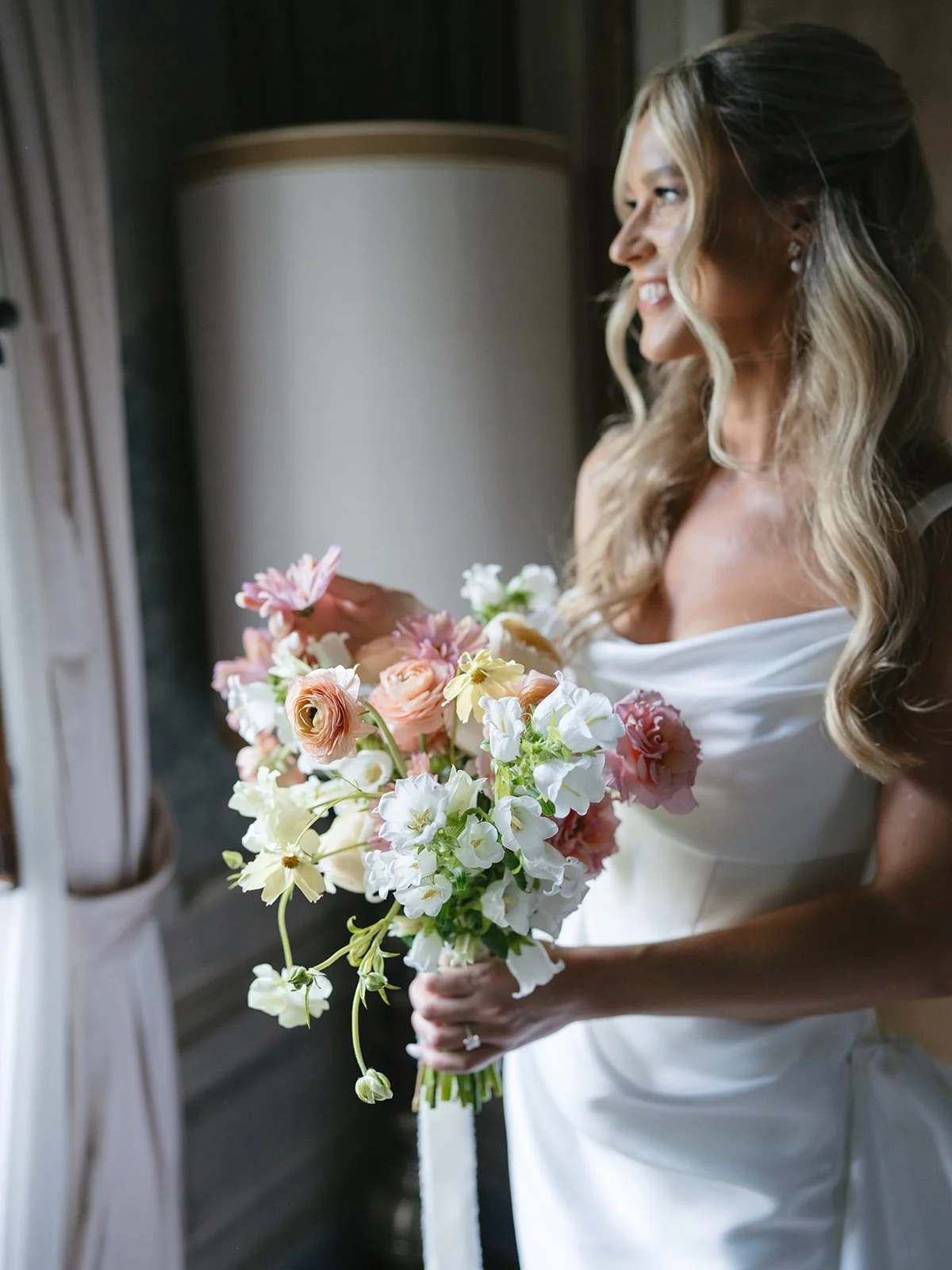 A bride in a white wedding dress holding a bouquet of pink, white, and cream flowers, smiling and looking out a window.