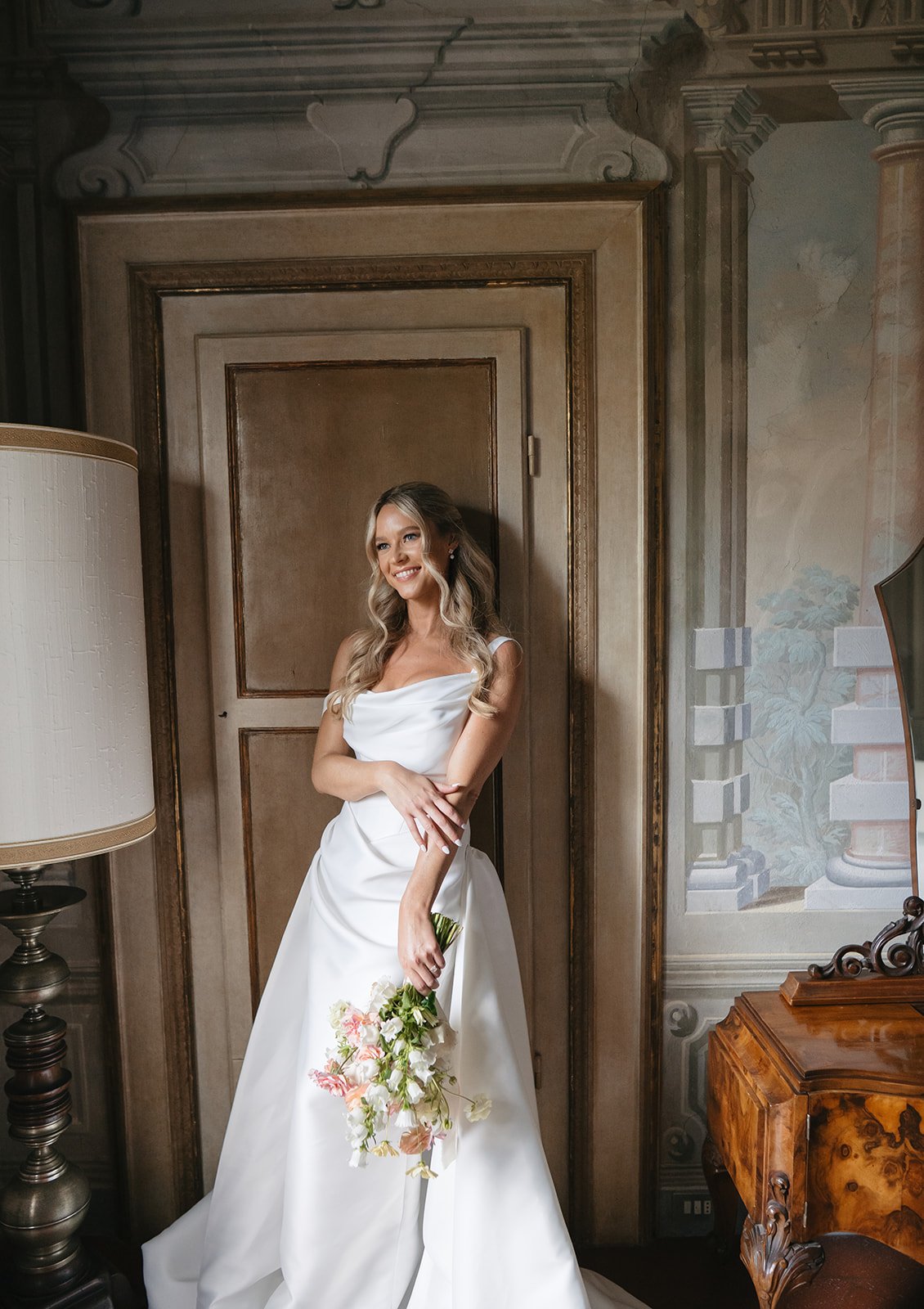 A woman in a white wedding dress holding a floral bouquet, standing inside a decorated room with ornate walls and furniture.