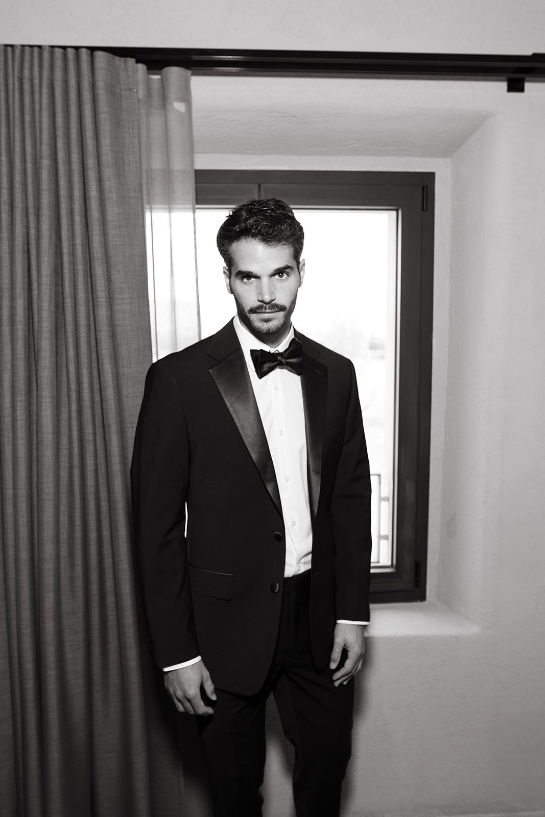 A man in a formal tuxedo and bow tie standing indoors in front of a window.