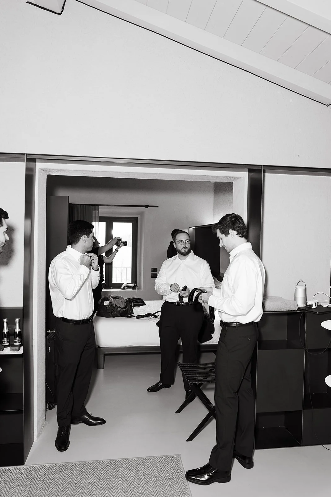 A group of men dressed in white shirts and black pants gathered in a room, with some holding cameras and a cellphone, preparing for a formal event or photoshoot, in a modern hotel room or suite.