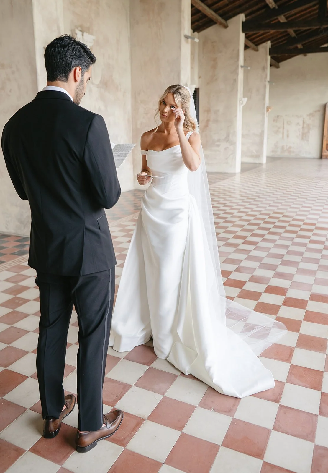 A bride in a white wedding gown and veil stands with an emotional expression, wiping away tears while holding a piece of paper, as a groom in a black suit reads from a paper during their wedding ceremony in an indoor venue with a checkered tile floor