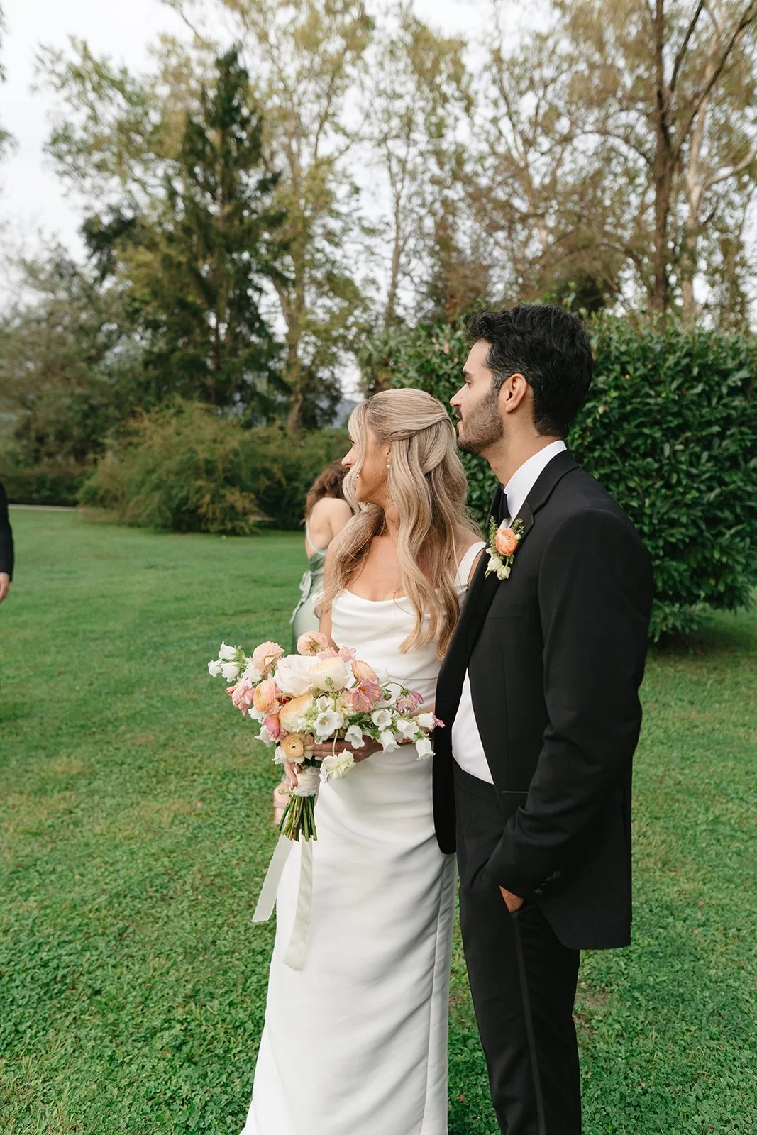 A bride and groom standing outdoors on a grassy area during their wedding ceremony, with the bride holding a bouquet of pink and white flowers.
