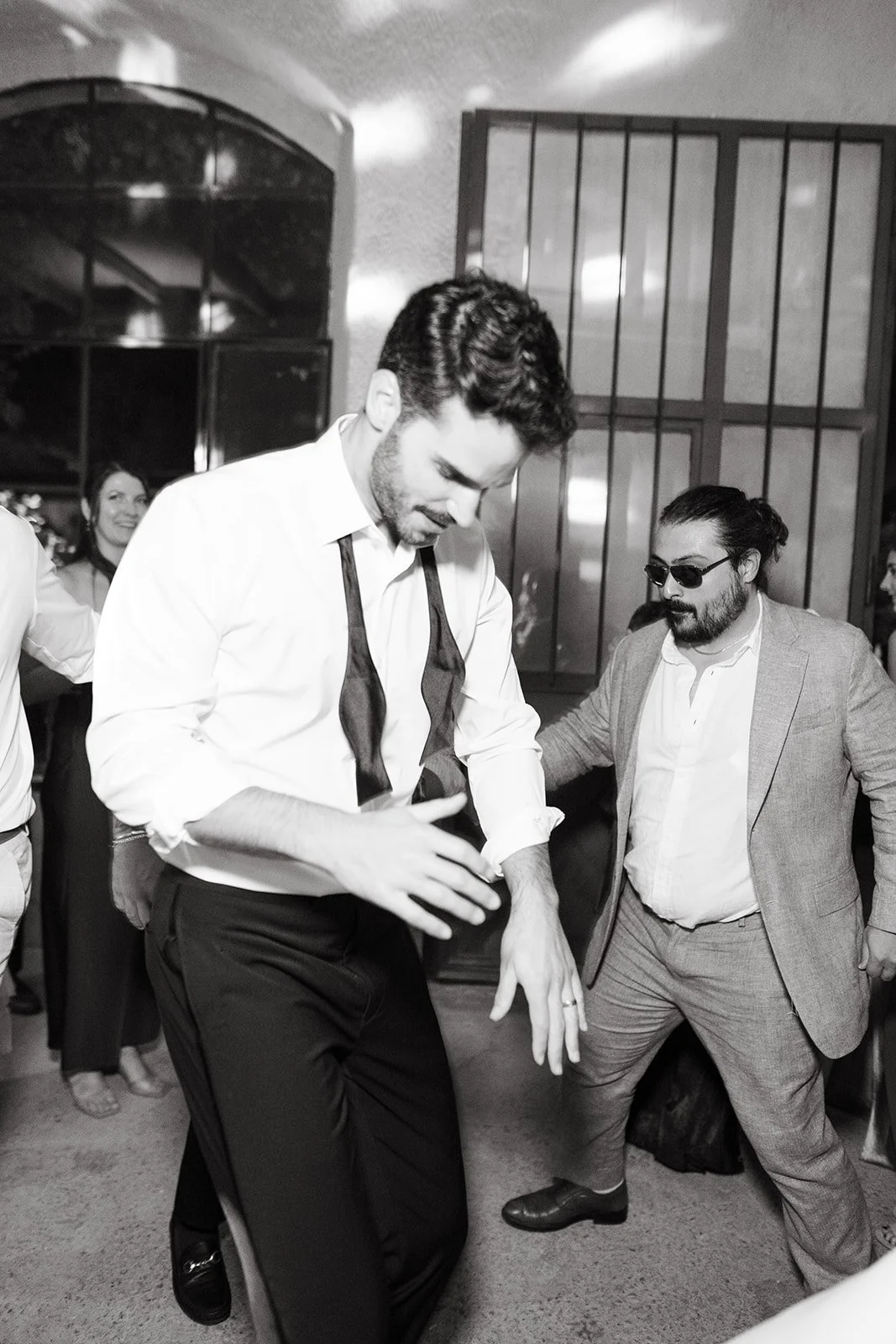 Two men dancing at an indoor party. The man on the left is dressed in a white shirt with rolled-up sleeves and a loosened tie, while the man on the right is wearing sunglasses, a light-colored blazer, and dress pants. People are visible in the backgr