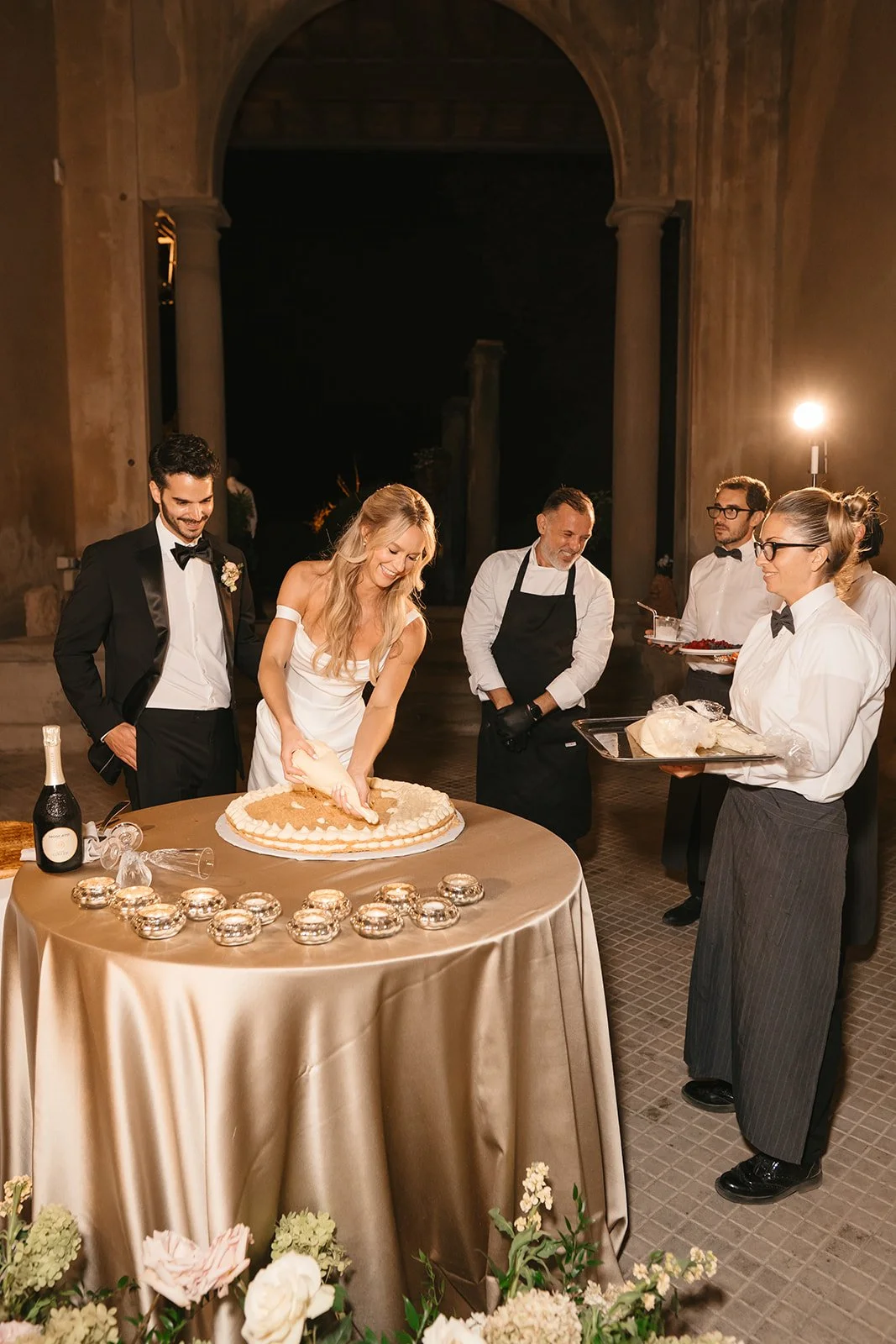 A woman, dressed in a white gown, cuts a wedding cake at her wedding celebration. She is surrounded by formally dressed guests and servers, with a golden tablecloth and floral decorations in the foreground.
