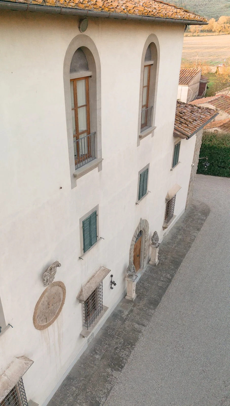 A tall, white building with tall arched windows and green shutters, situated on a narrow street with stone pavement and a rural landscape in the background.