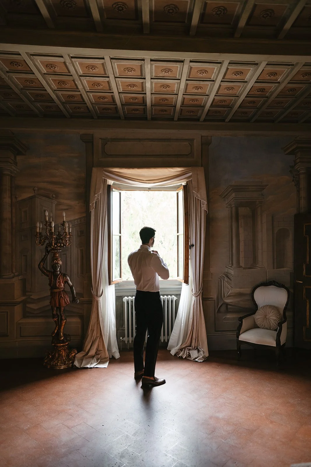 A man dressed in formal attire standing in a vintage room with elegant furniture and artwork, looking out a large open window with sunlight streaming in.