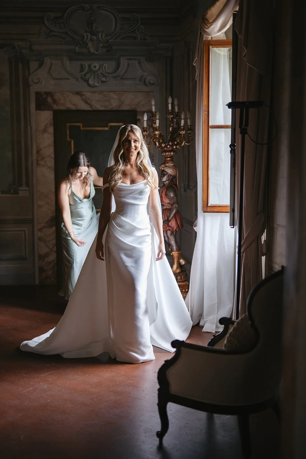 Bride in a white wedding dress smiling with a bridesmaid in a green dress adjusting her gown in an elegant room with ornate wall decorations and a window letting in natural light.