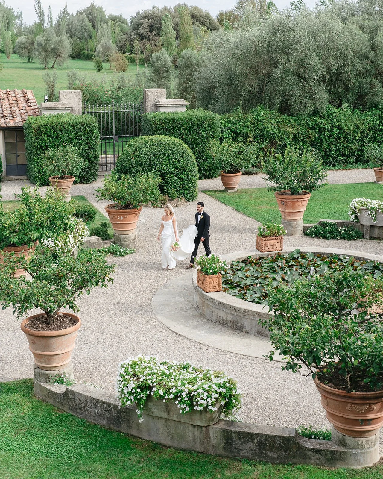 Bride and groom walking together in a lush garden, surrounded by potted plants, trees, and greenery.
