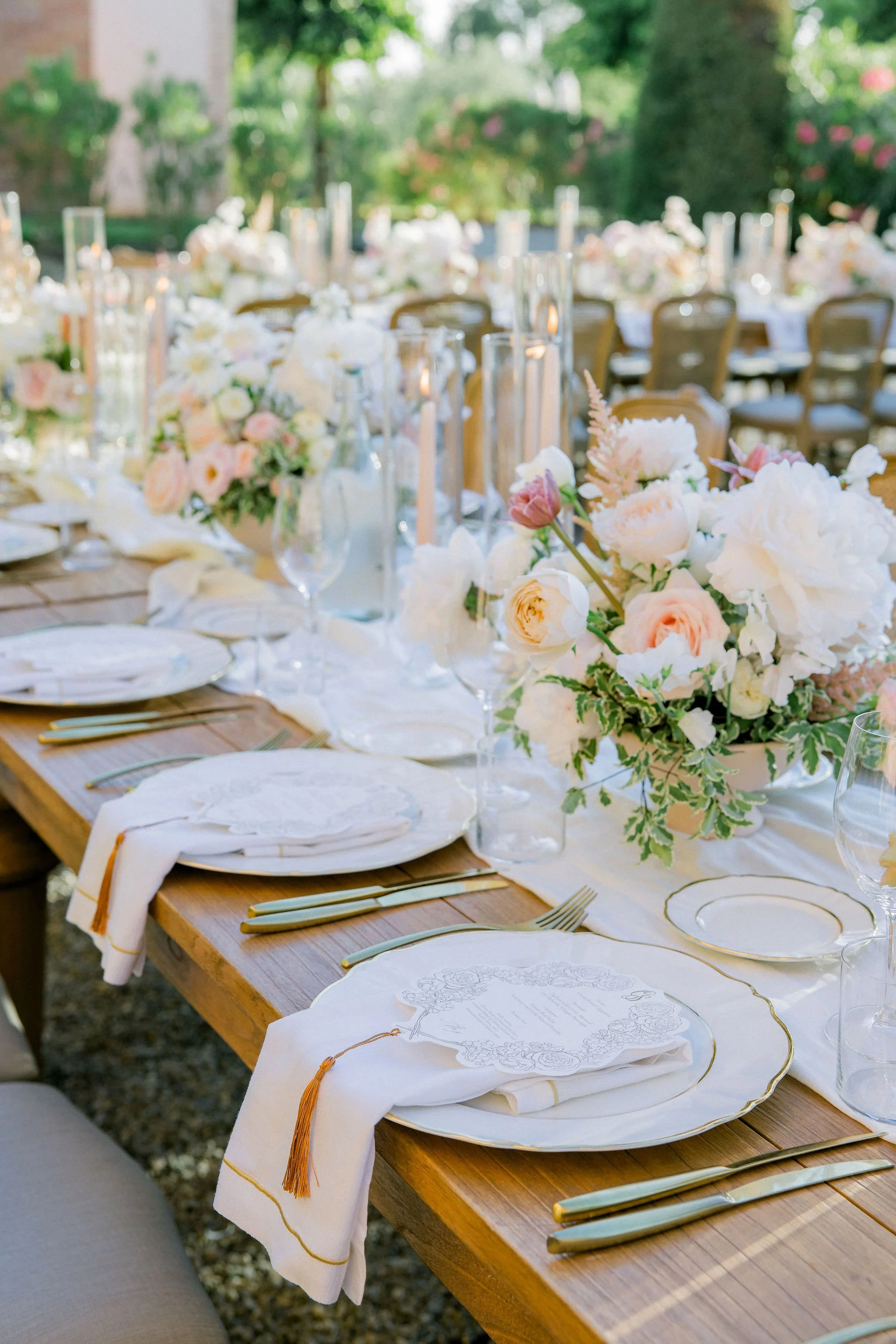 Elegant outdoor wedding table setting with floral centerpieces, candles, plates, and gold cutlery.