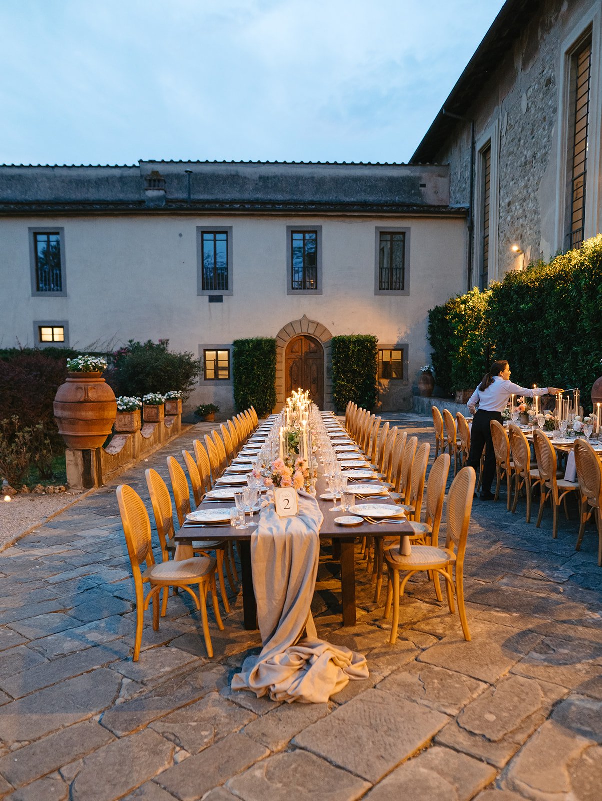 Long dining table set outdoors in a courtyard with a stone floor, decorated with floral arrangements and candles for an evening event, with a large building in the background and a server attending.
