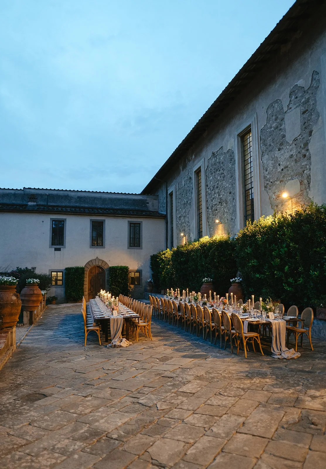 Outdoor wedding or dinner setup with long tables decorated with candles, surrounded by arched and rectangular windows on stone and stucco walls, under evening sky.