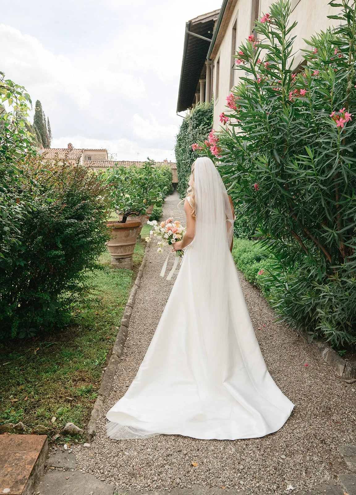 A bride in a white wedding dress with a veil, holding a bouquet, walking along a garden path surrounded by green plants and pink flowers.