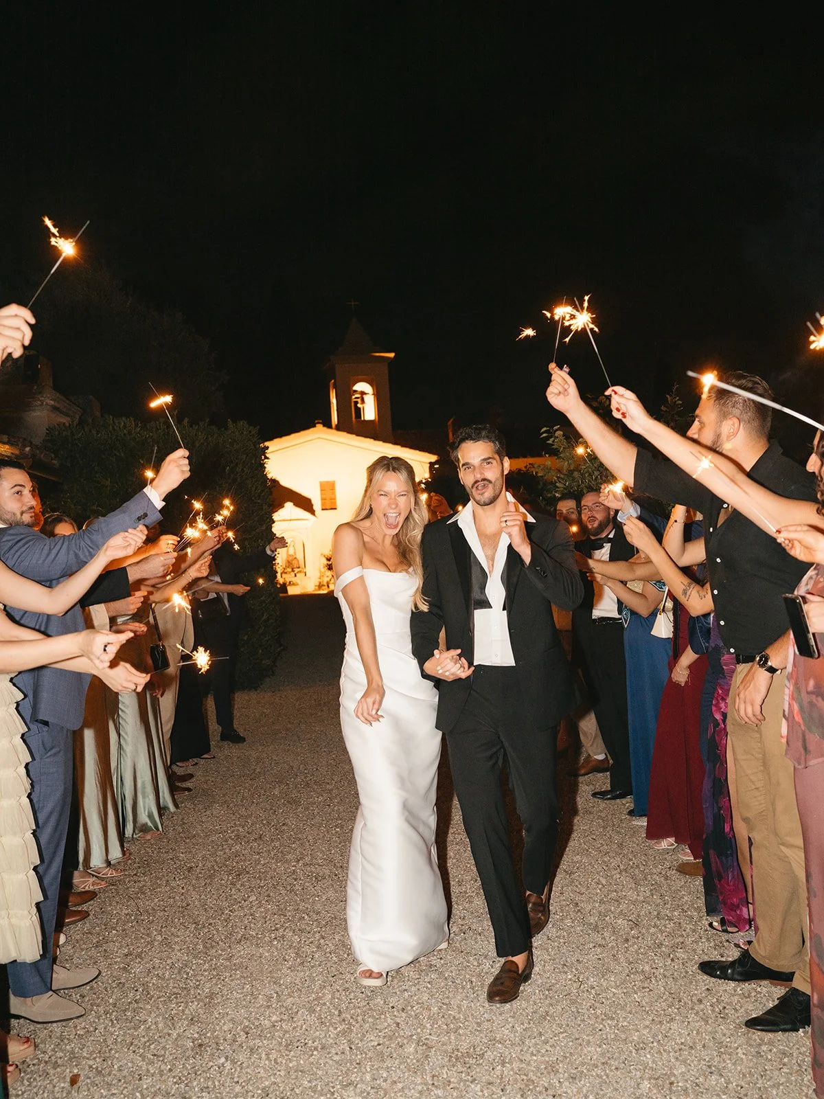 Couple in wedding attire walking through a night celebration, surrounded by guests holding sparklers in an outdoor setting near a lit building.