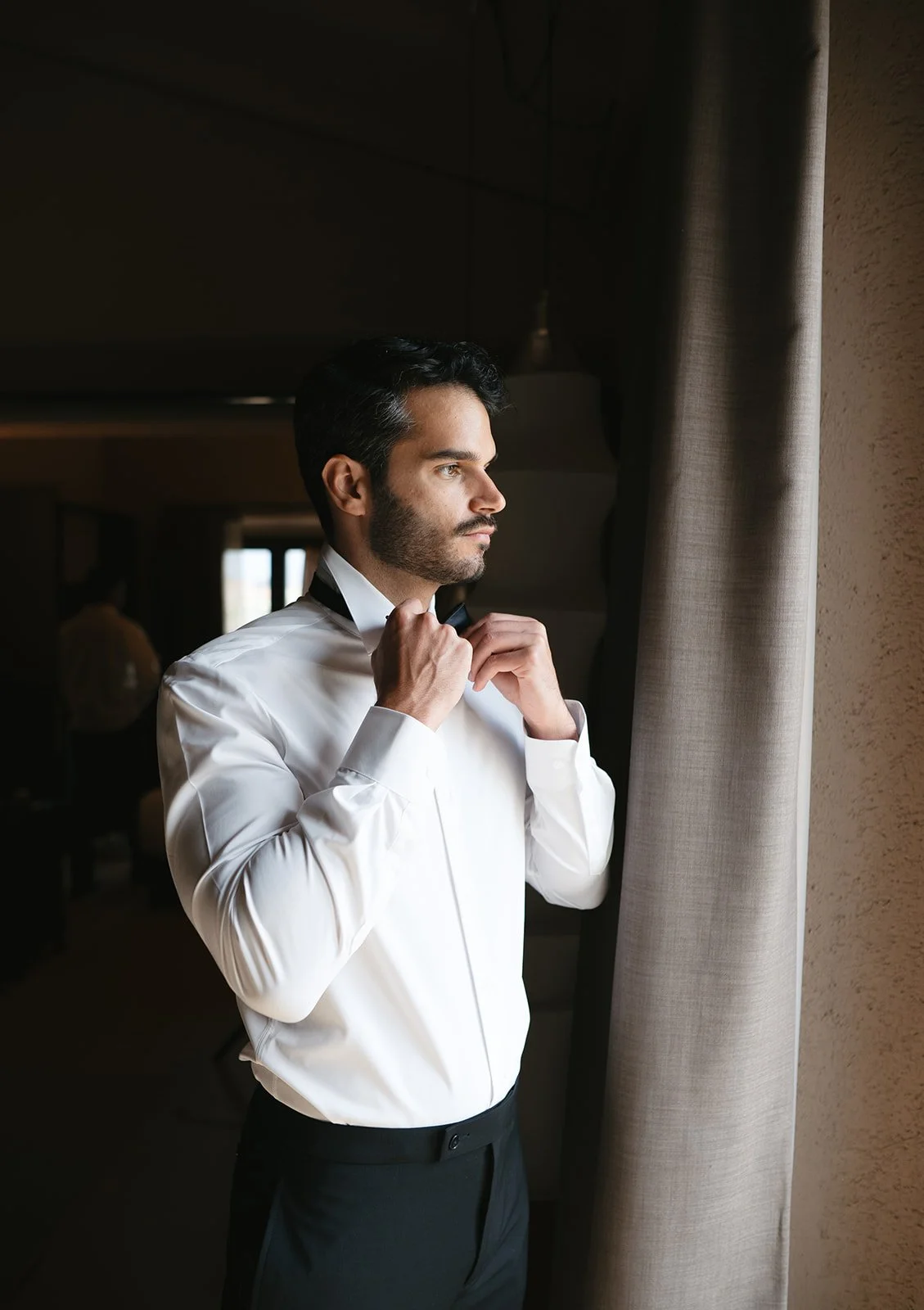 A man in a white dress shirt adjusts his bow tie near a window, preparing for an event.