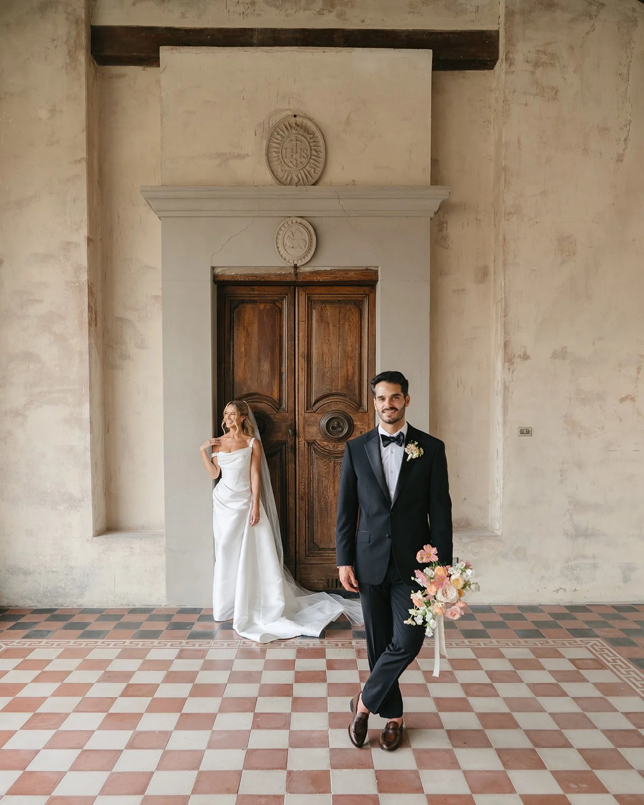 A bride and groom in a historic building with high ceilings, textured walls, and brick flooring. The bride, in a white gown with a veil, stands in the background near a wooden door. The groom, in a black tuxedo with a bow tie, stands in the foregroun
