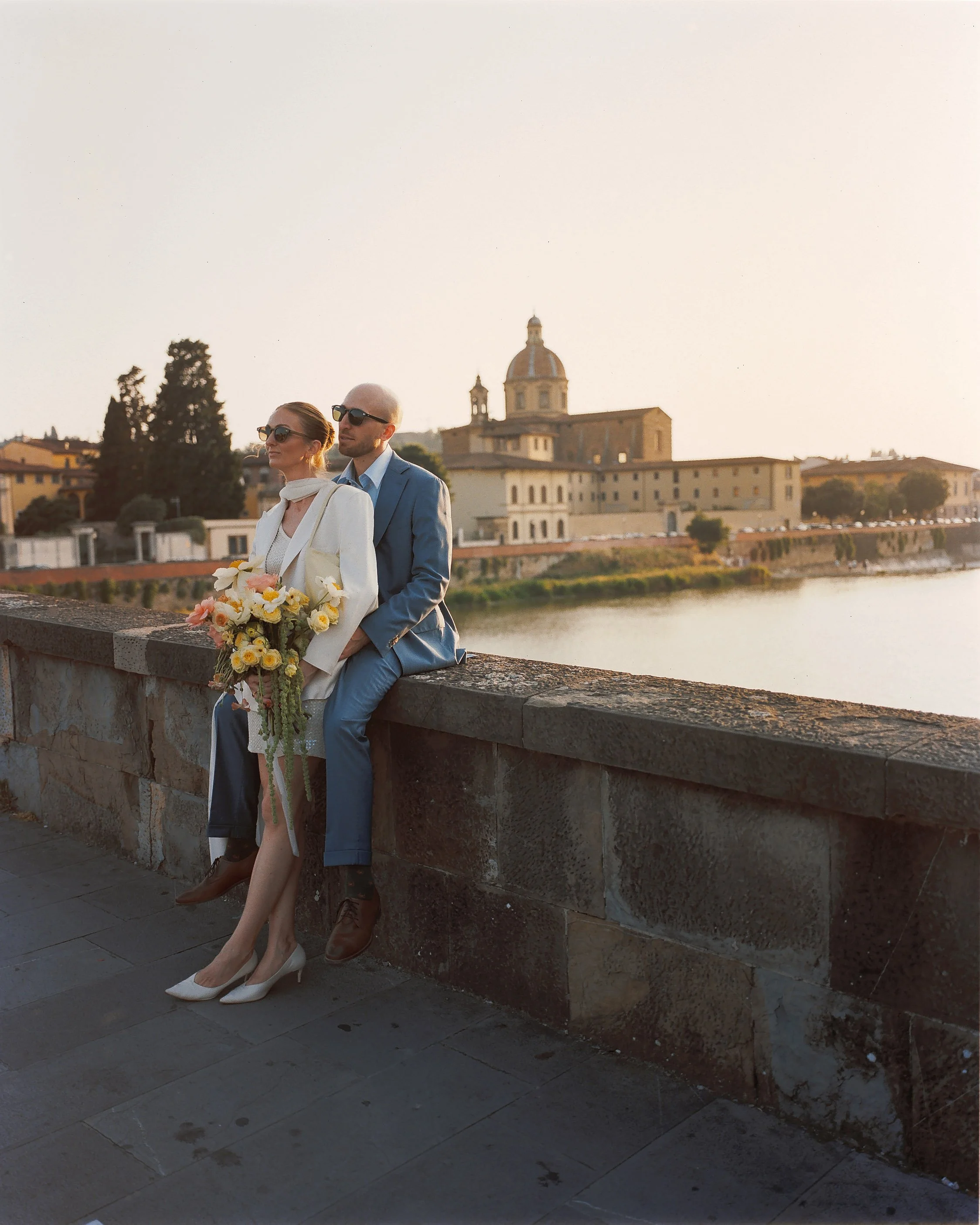 A couple standing in a garden overlooking a cityscape, with the man pointing towards a landmark in the distance.