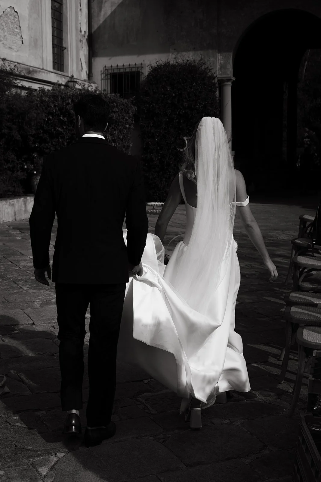 A bride in a white wedding dress and veil, holding her dress, walking with a groom in a black suit, in an outdoor courtyard with chairs on the side.