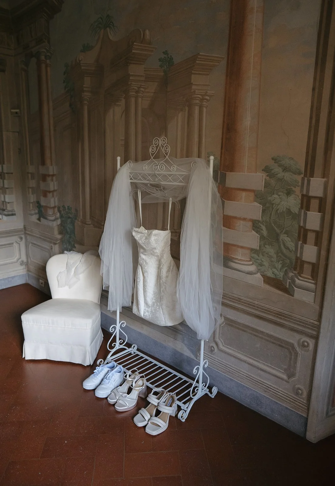 Wedding dress hanging on a white decorative rack with a tulle veil, white sneakers, and white high-heeled shoes below. A white upholstered chair is next to the rack, and a mural with architectural columns and greenery is in the background.