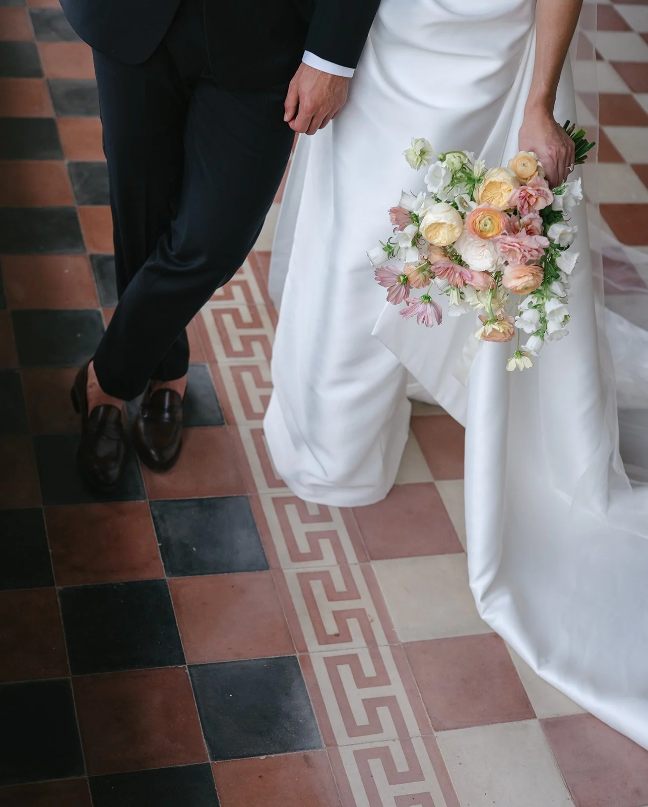 A bride and groom standing side by side on a patterned tile floor. The bride is holding a bouquet of pink, white, and peach flowers, and her white wedding dress is draped to the floor. The groom is wearing a black suit with loafers.