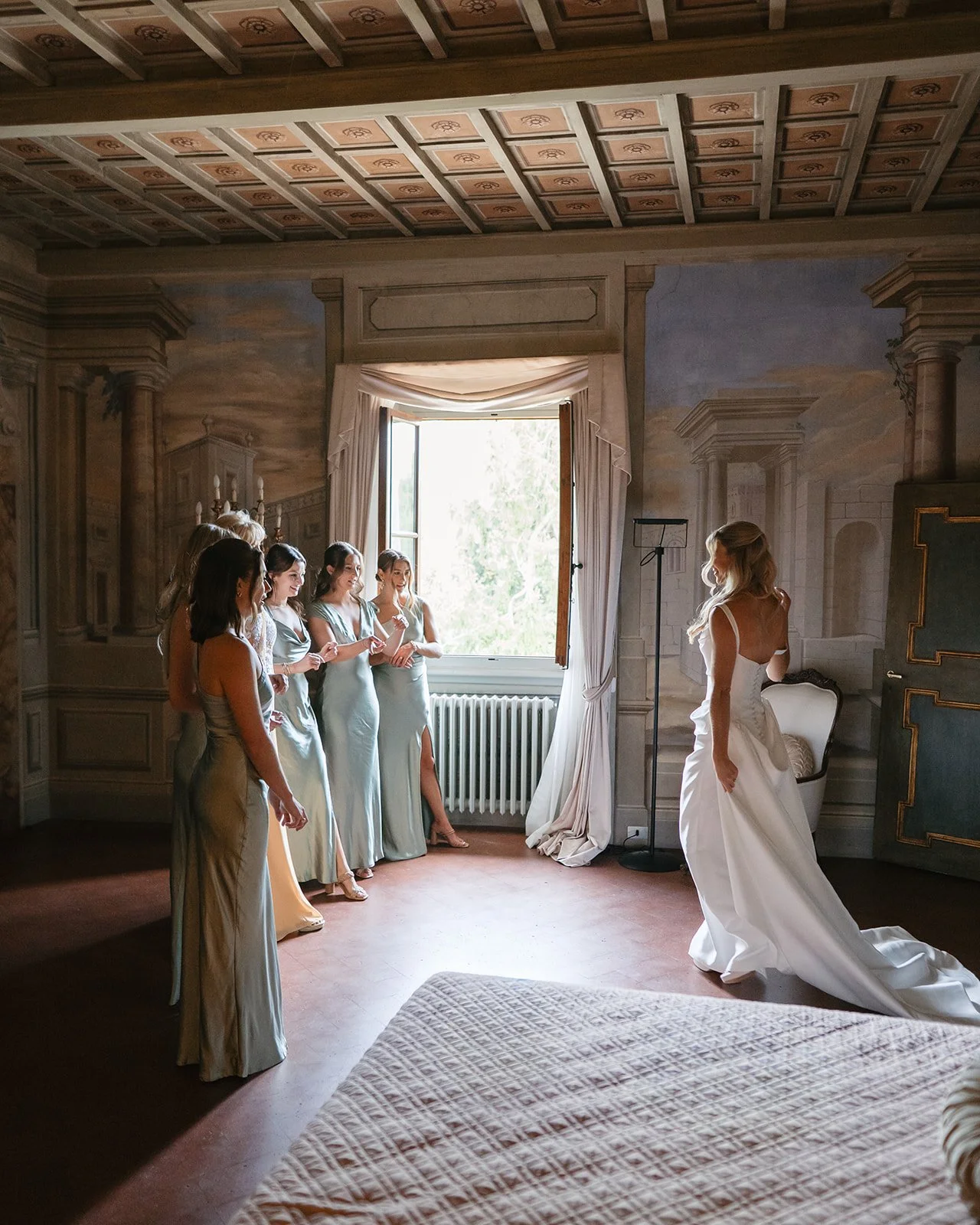 Bridesmaids and bride in a room with ornate wall murals and large window, with the bride in a white gown and the bridesmaids in light-colored dresses.