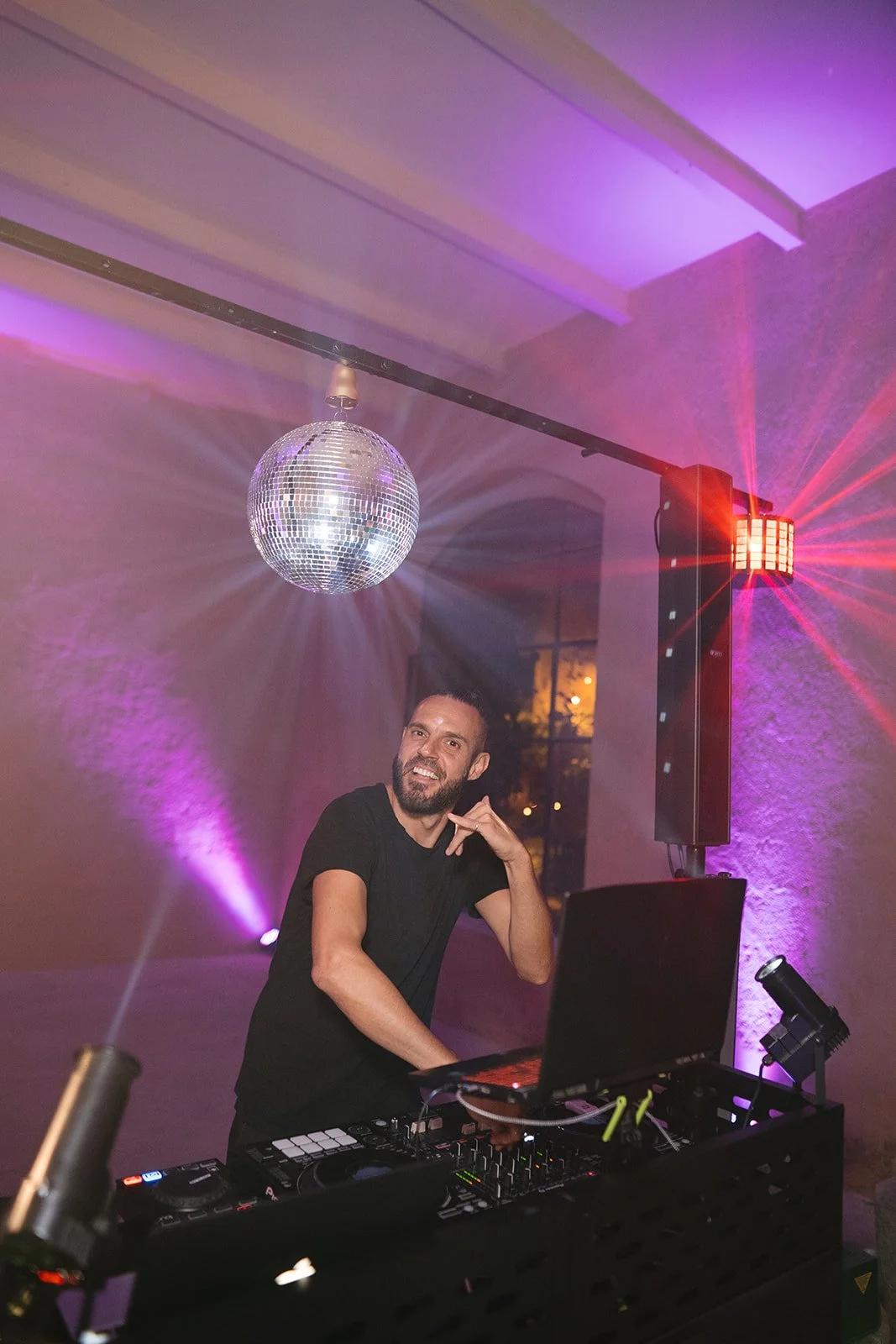 A DJ with a beard and black shirt smiling and making a gesture with his hand while standing behind a DJ booth at a party, with colorful lighting and a disco ball above him.