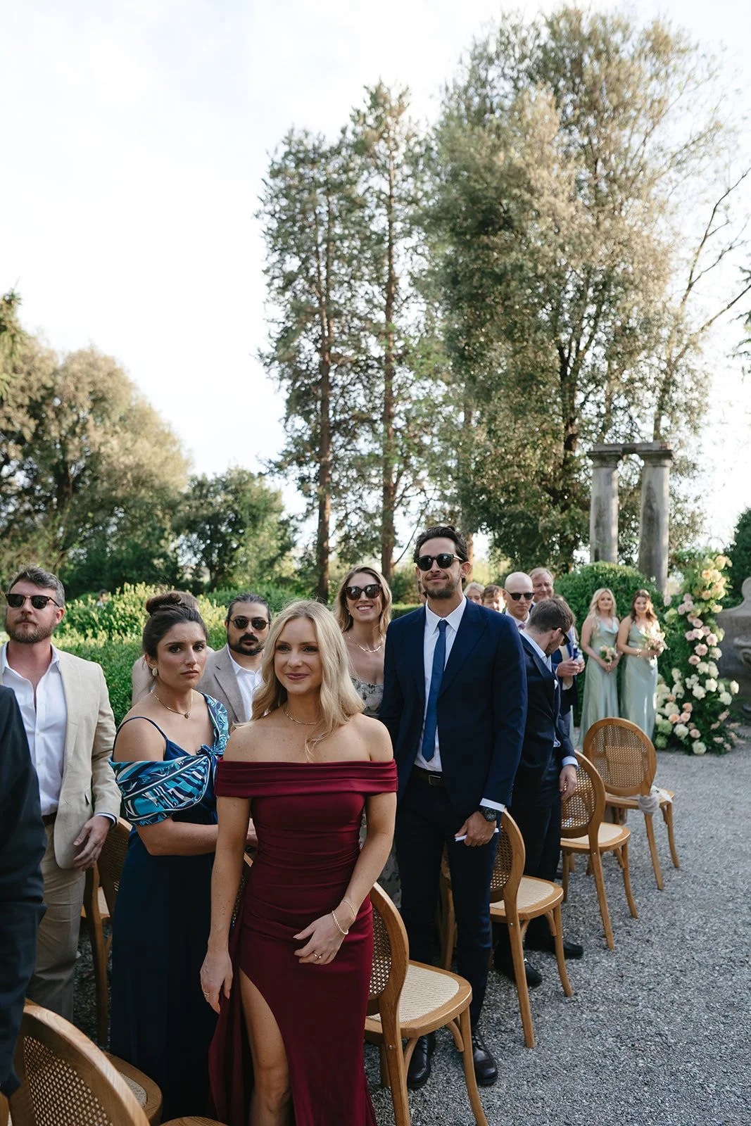Group of people dressed in formal attire attending an outdoor wedding or event, with trees and floral decorations in the background.
