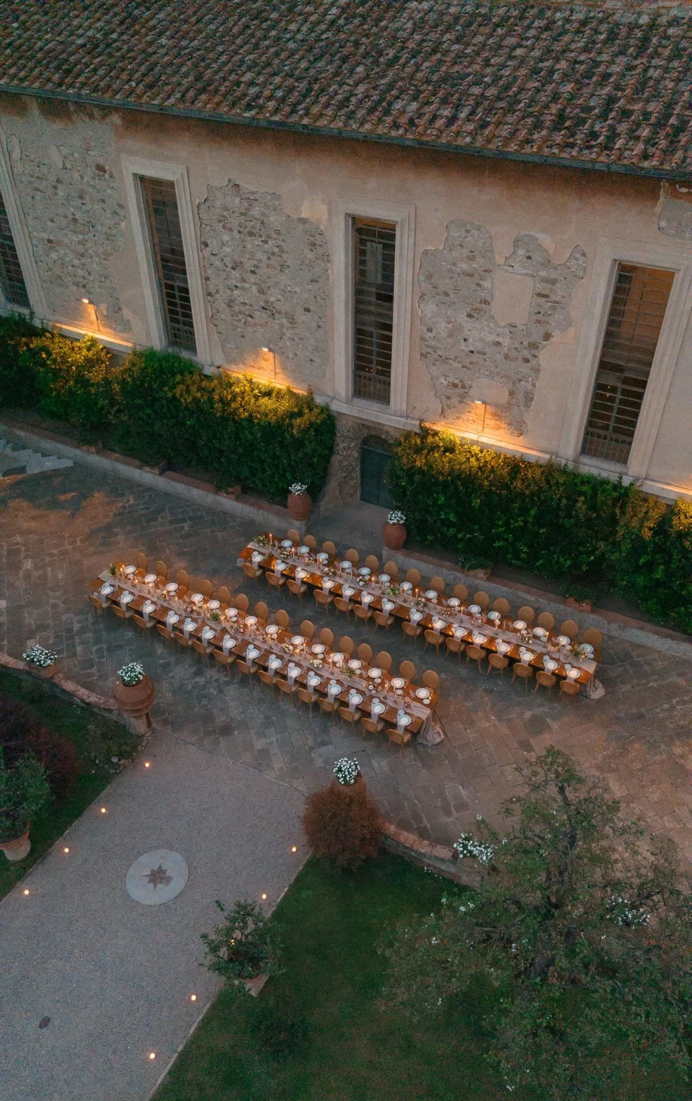 Outdoor evening event setup with long banquet tables arranged on a stone patio, decorated with tableware and flowers, near a historic stone building with worn walls and tall windows, surrounded by greenery and illuminated by soft lighting.