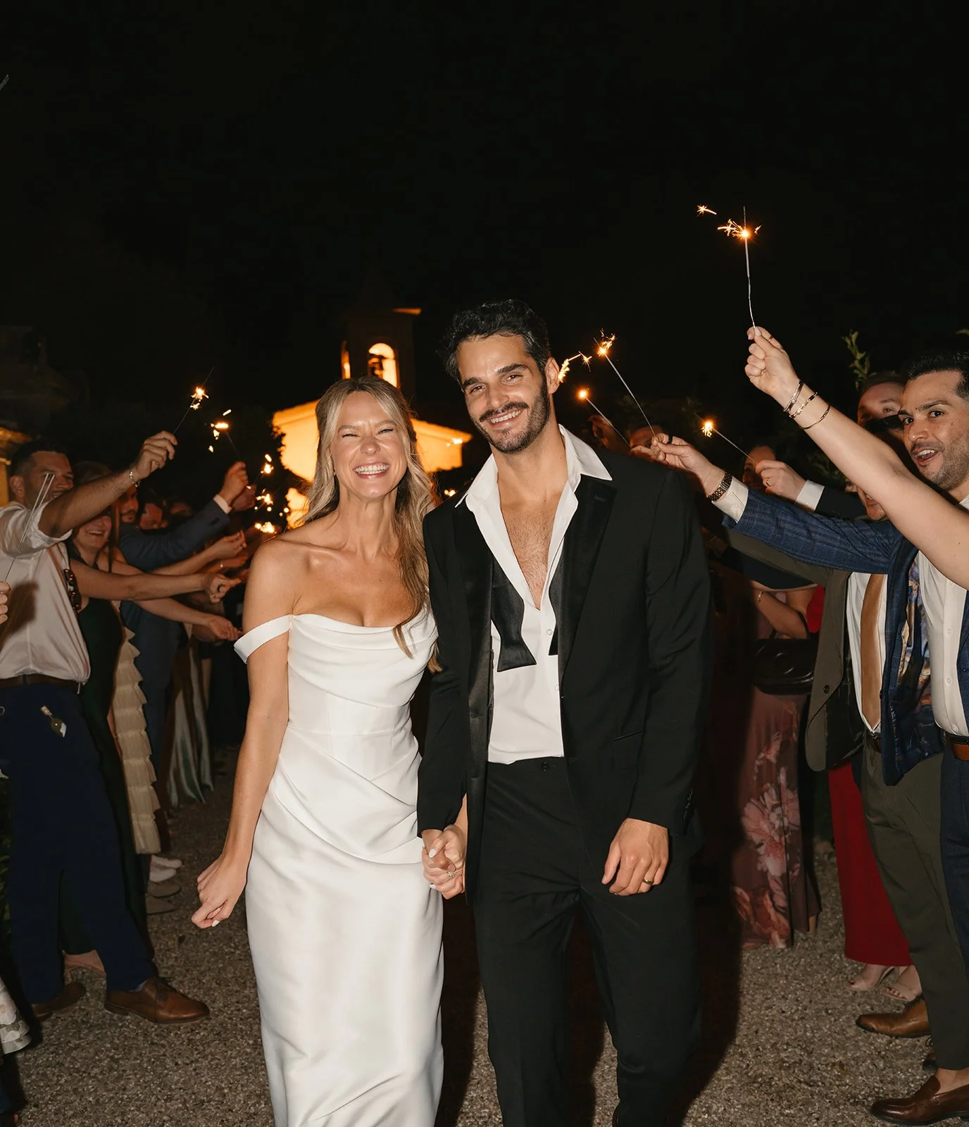 A couple dressed in formal wedding attire holding hands and smiling at night, surrounded by guests holding sparklers.