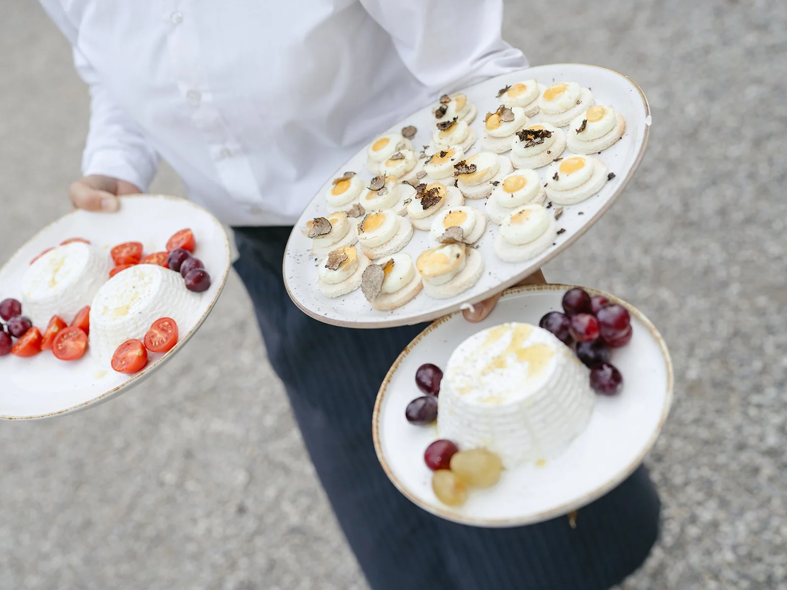 Person holding three plates of assorted desserts, including mini cupcakes topped with chocolate and truffles, panna cotta with berries, and a dome-shaped dessert with yellow sauce and berries.