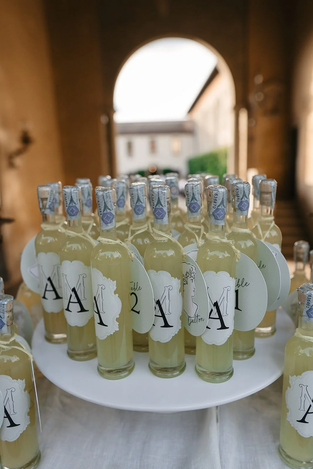 Multiple glass bottles of a light yellow beverage on a white table, each with a label and sealed with a blue foil cap. The labels feature a black monogram A and the number 1 or 2, indicating table assignments, with some labels having a line drawing o